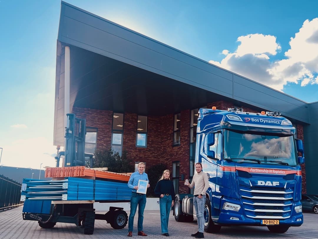 Blauwe vrachtwagen en oplegger met de achtergrond een blauwe loods en drie mensen met een certificaat